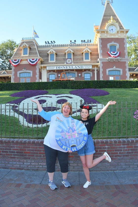 Two women, Cami and her mother, stand in front of a building and Mickey Mouse-shaped garden with a circle sign that reads "We're 64 today!" for Disneyland's 64th anniversary. 