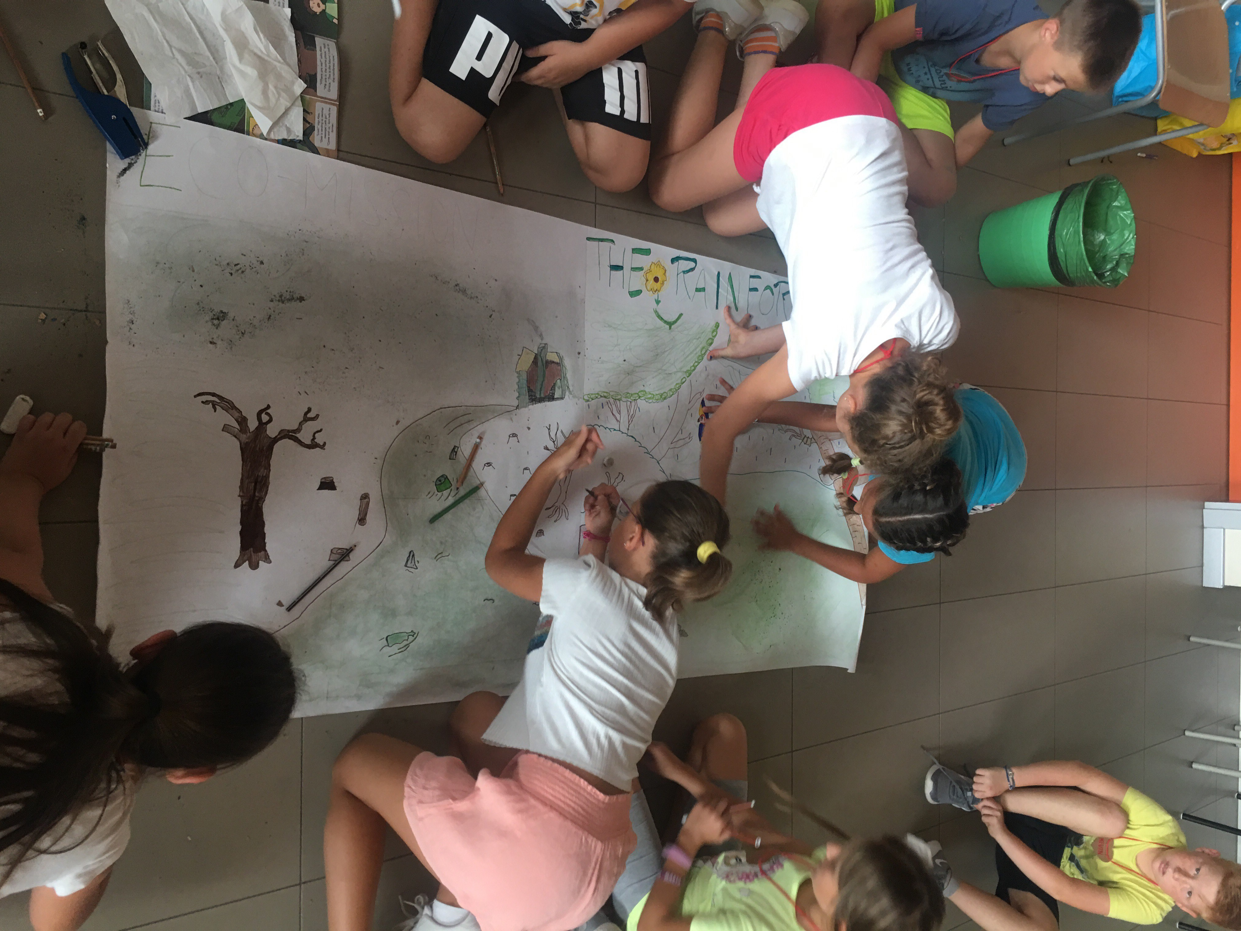 Several young children lean over a large sheet of paper, coloring a landscape.