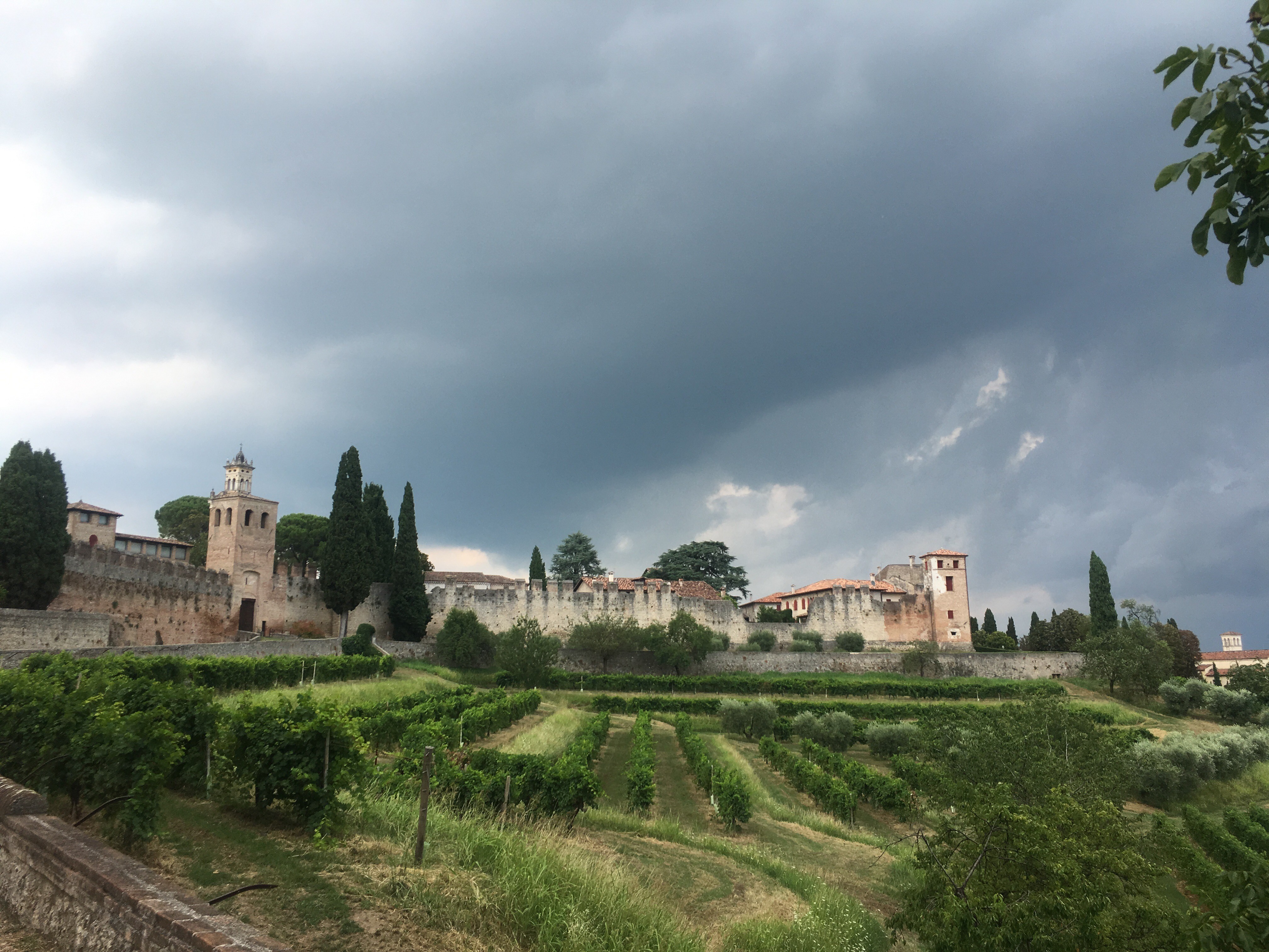 A beautiful Italian landscape with old, white buildings in the background, rolling green hills in the foreground, and stormy clouds brewing.