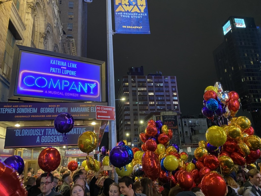Photograph is of a crowd gathered for the preview of the show "Company" with the theater sign glowing bright blue in the background. Many people hold colorful balloons.