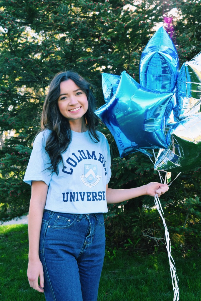 Image shows a young woman with long dark hair wearing a blue shirt and jeans and holding blue and silver star-shaped balloons. The shirt reads "Columbia University" and shows the university seal. In the background is a dark spruce tree on a sunny day.